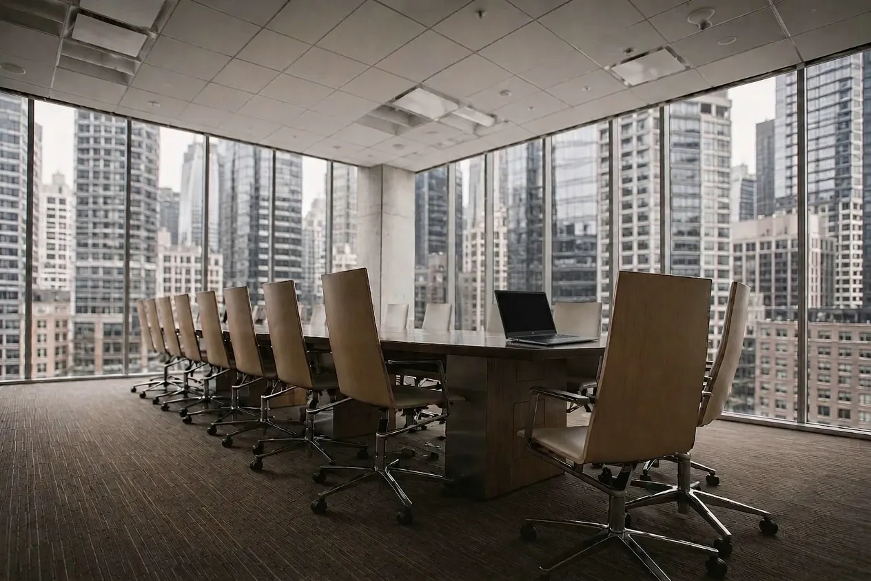 An empty boardroom high above a city, chairs drawn up to a long table and a single laptop open - the room where steering committee decisions are made